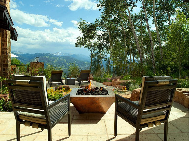 A full shot of four chairs and a bonfire with trees and a mountain view.