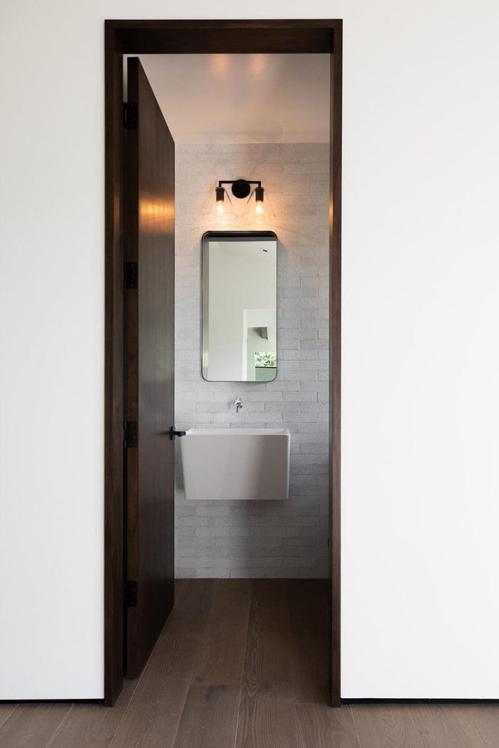 A full shot of a brown comfort room door, a white wall, and a wooden floor Light bulbs, a comfort room sink, and a mirror.